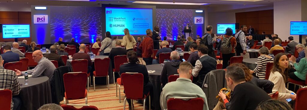 Delegates of PCI London at tables in front of a display showing event sponsors including Ground Labs